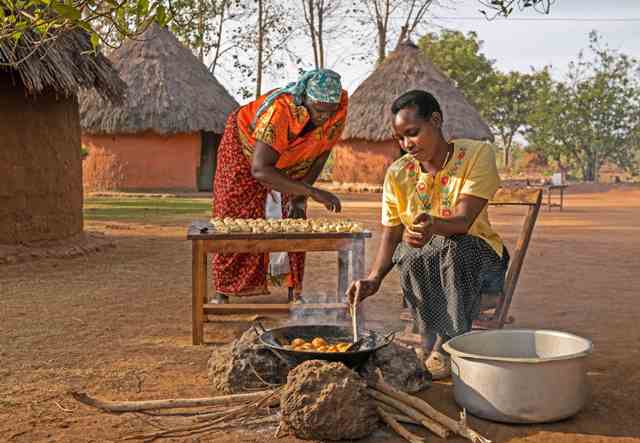 Women cooking outdoors in rural East Africa with traditional huts in background.