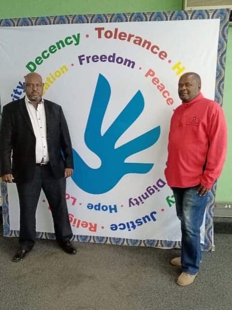 Two men standing beside a colorful banner emphasizing values like decency, freedom, and peace.