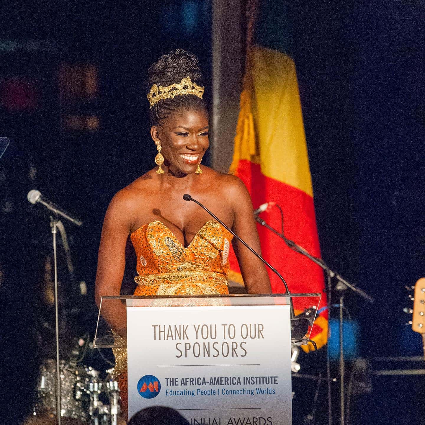 African woman speaking at awards event with African flag in background.