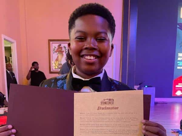 Young African boy in a tuxedo holding a certificate at a cultural awards ceremony.