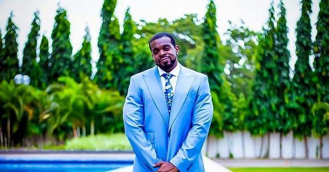 Pryme in a blue suit standing outdoors with lush greenery background.