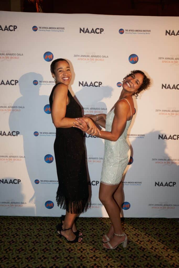 Two women smiling and holding hands at the NAACP Awards Gala, showcasing African culture and unity.