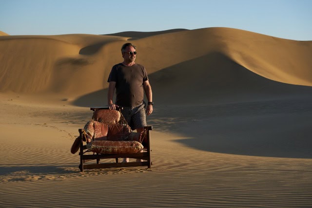 Man standing in desert with sofa and luggage, dunes in background.