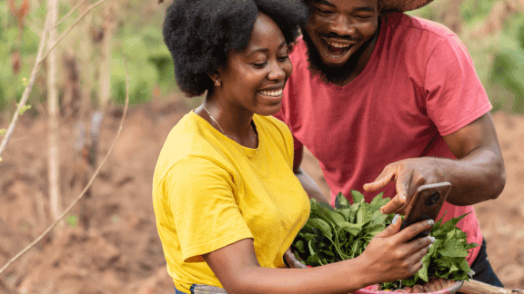 Two Kenyan women farmers examining crops and using a mobile phone outdoors.