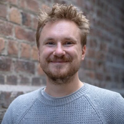 About Us 18 Smiling man with curly hair and beard standing against a brick wall.