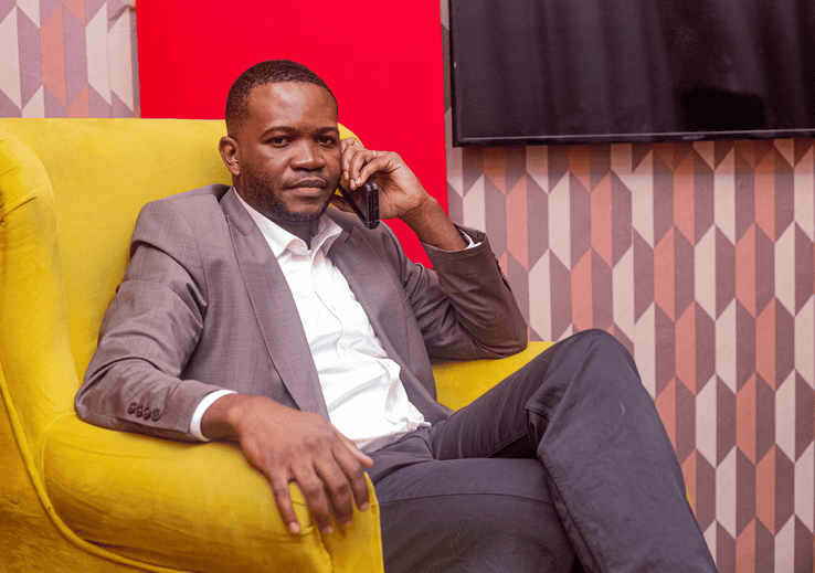 Man in suit sitting on yellow sofa, talking on phone, in an office setting.