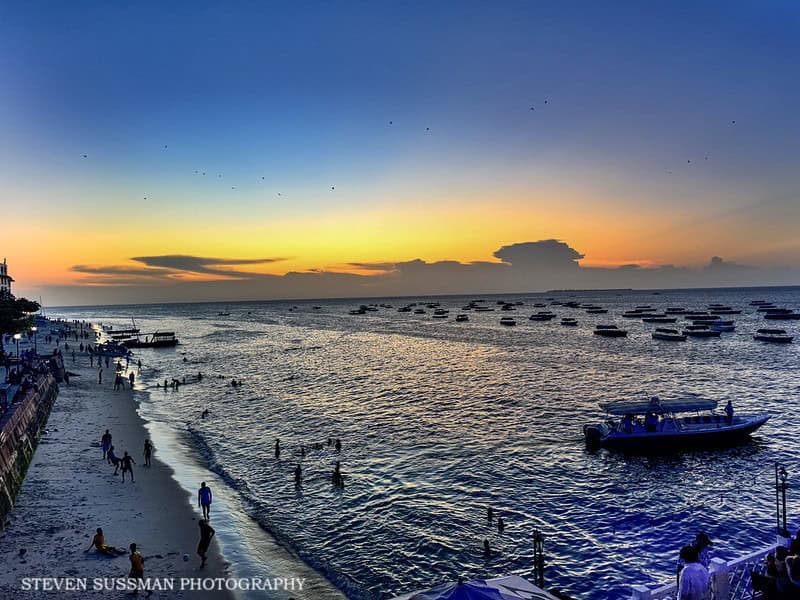 Sunset over Zanzibar beach with boats, people, and vibrant sky colors.