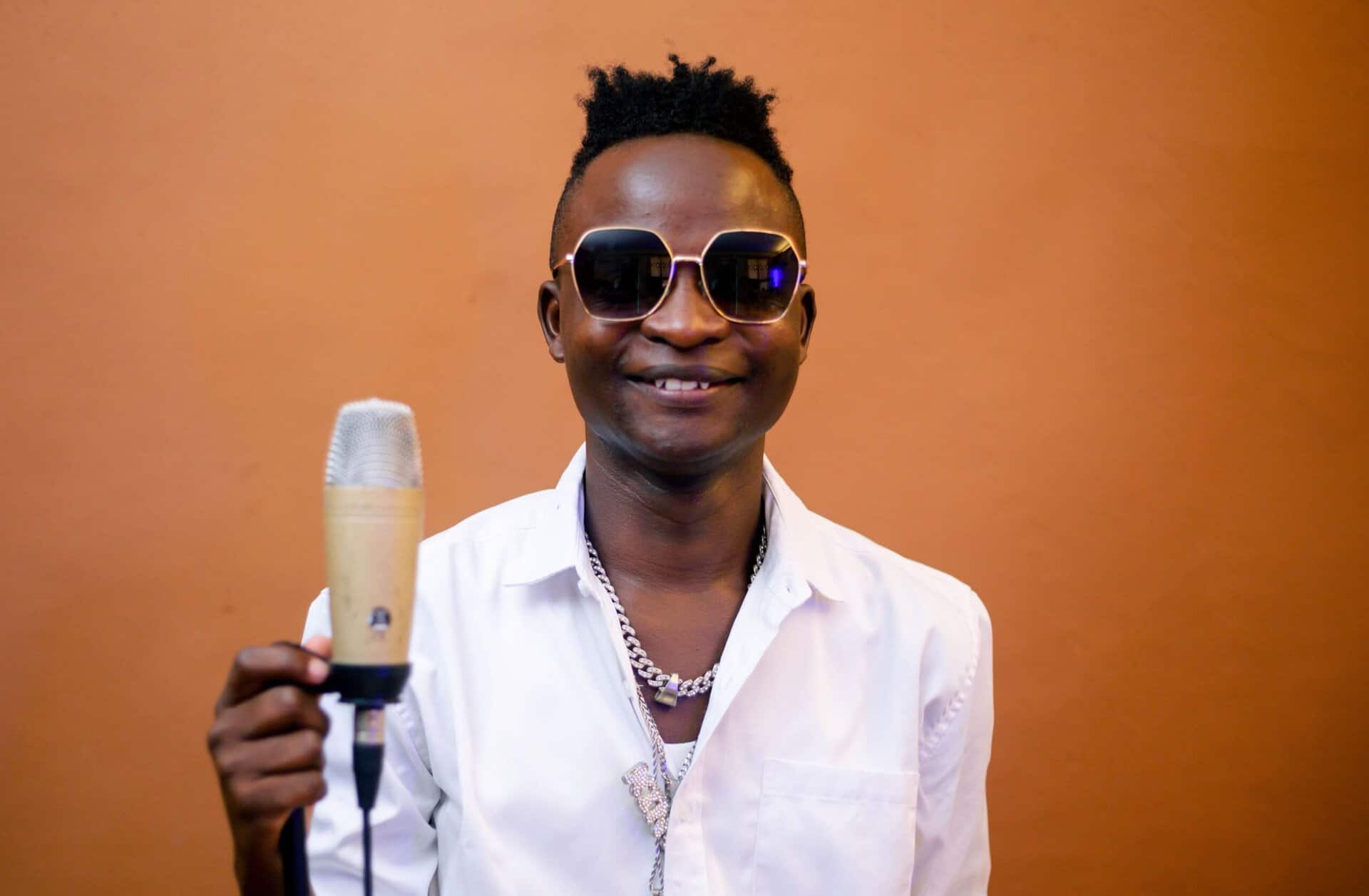 Gabiro Mtu Necessary holding a microphone, smiling, wearing sunglasses and a white shirt.