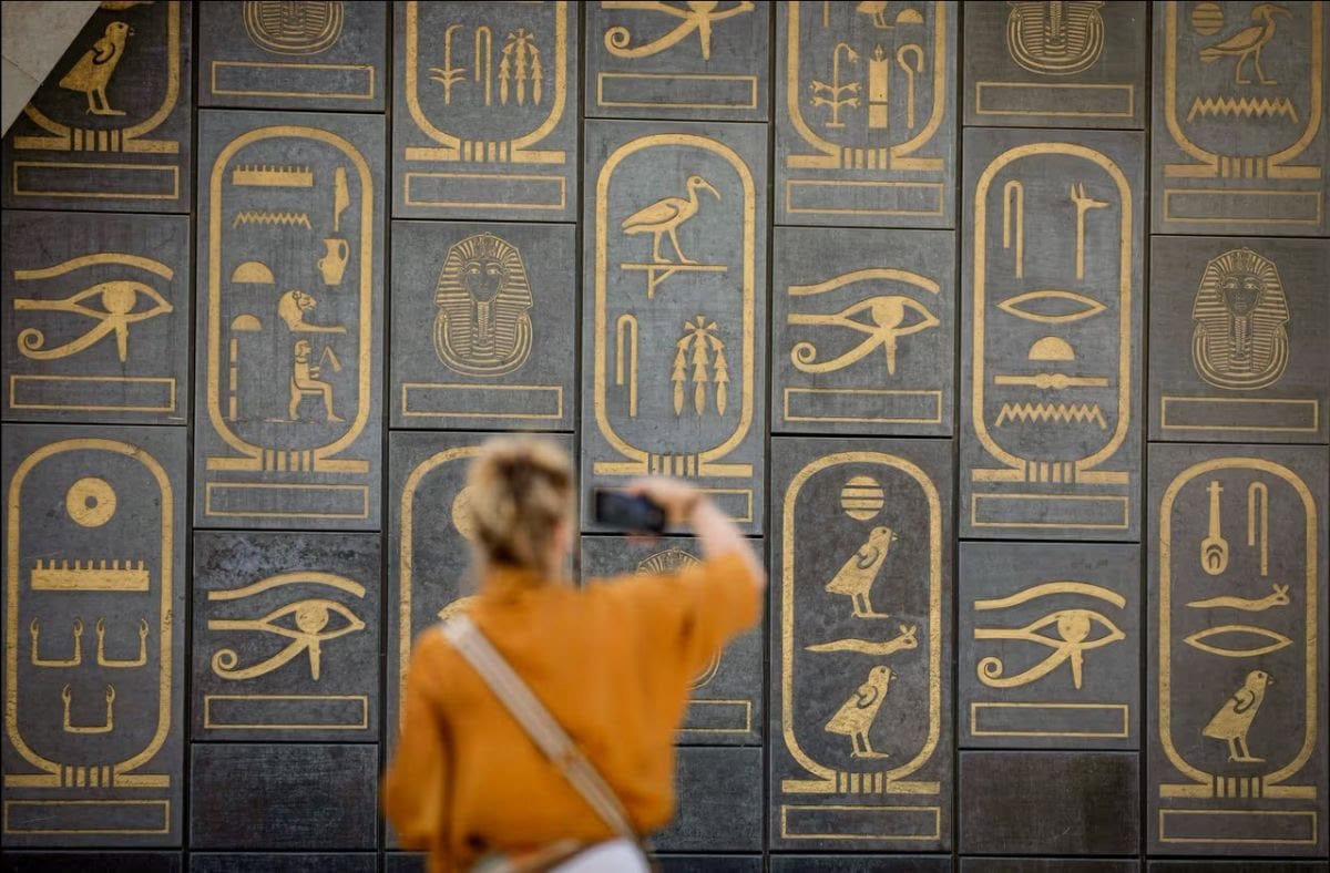 Person photographing ancient Egyptian hieroglyphs on museum wall.