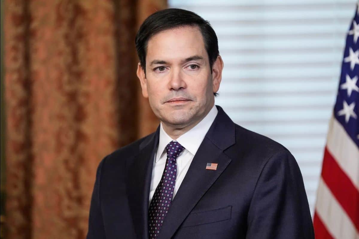 U.S. politician in a dark suit and tie, standing in front of an American flag.