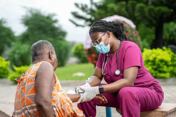Healthcare worker in pink scrubs and mask providing medical care to an elderly woman outdoors.