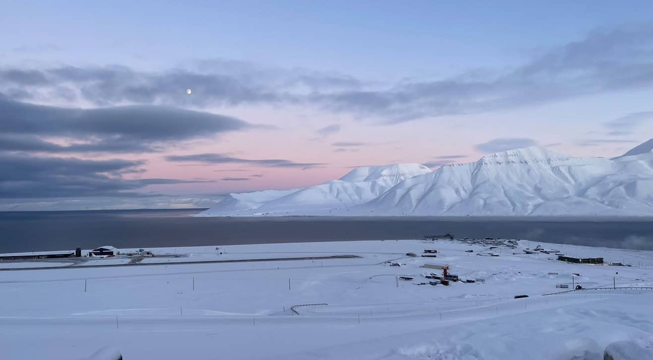 Snow-covered Arctic landscape with mountains and a calm sea at sunset.