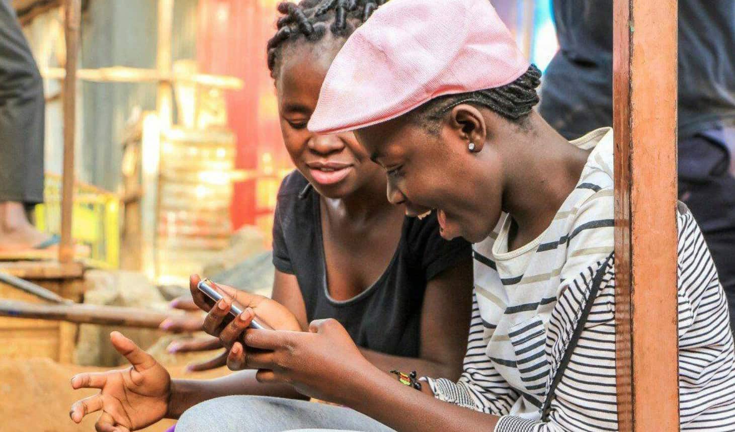 Two African women smiling and looking at a smartphone outdoors.