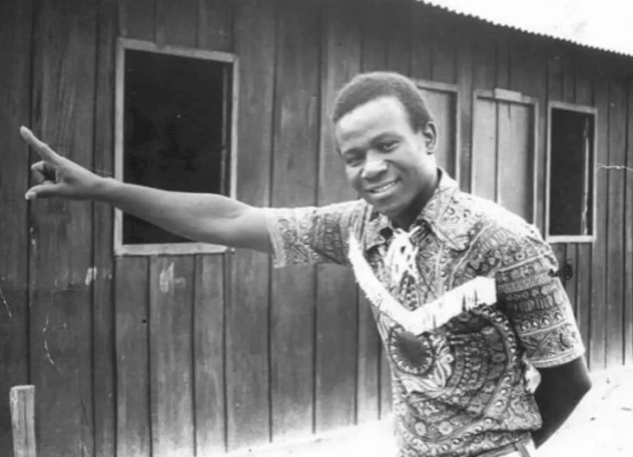 African man pointing at a wooden building in a rural setting.