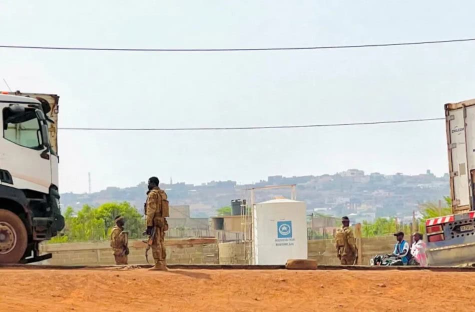 African soldiers patrolling on a dusty road with trucks and buildings in the background.