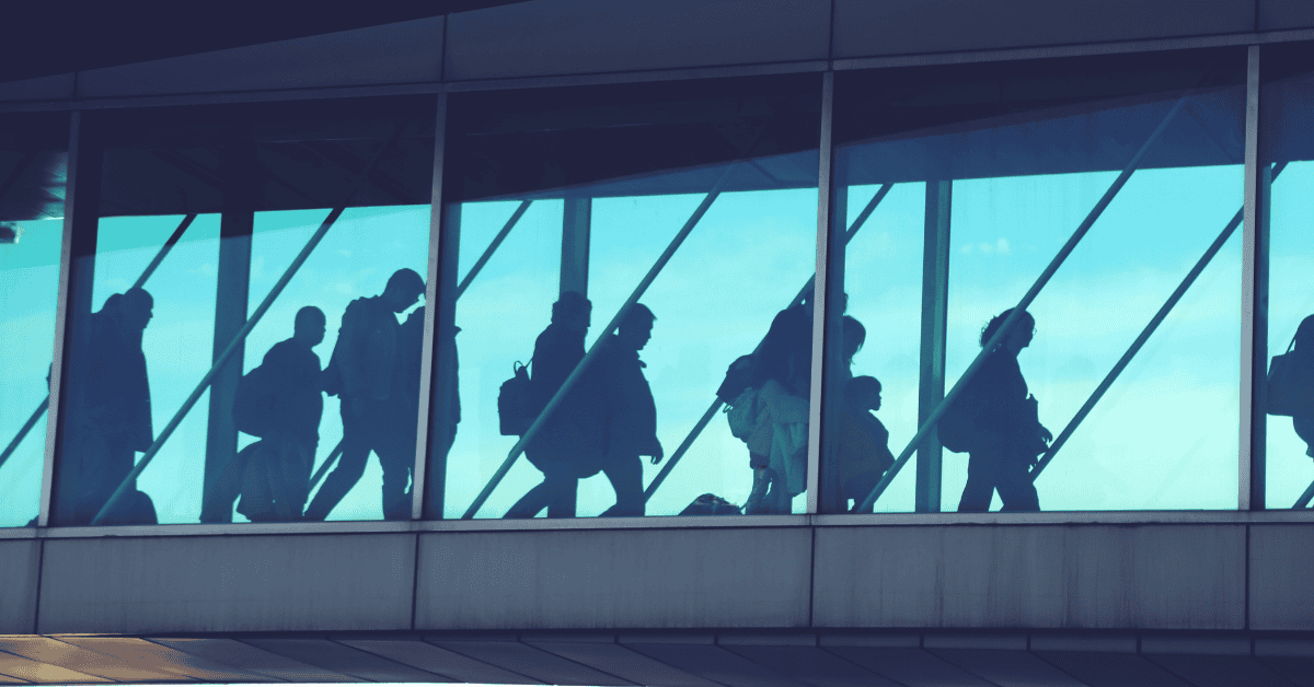 Refugees and migrants walking through an airport terminal with large glass windows.