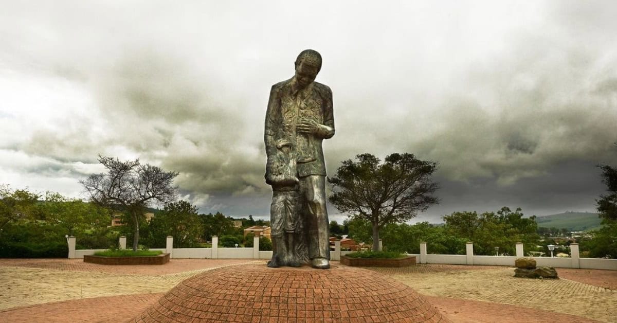 Statue of a man reading a book in an outdoor park with trees and cloudy sky in the background.
