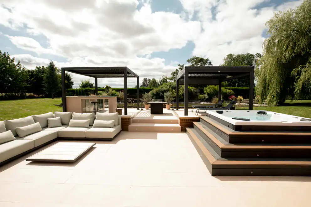 Luxury outdoor living space featuring two modern black pergolas with adjustable louvered roofs, creating shaded areas over stylish outdoor furniture including sectional seating and a hot tub, set in a beautifully landscaped garden setting