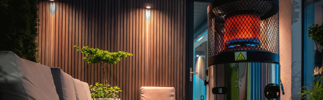 A modern outdoor heater emitting warm light, set against a wooden wall with greenery in the background.