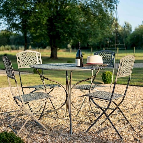 Metal bistro garden set with four folding chairs and a round table, placed on gravel in an outdoor setting. A wine bottle, glass, and straw hat sit on the table, with trees and open countryside in the background.