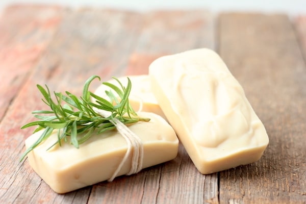 Image shows three bars of rosemary mint goat milk soap on a table. One has a sprig of rosemary tied around it with white string.