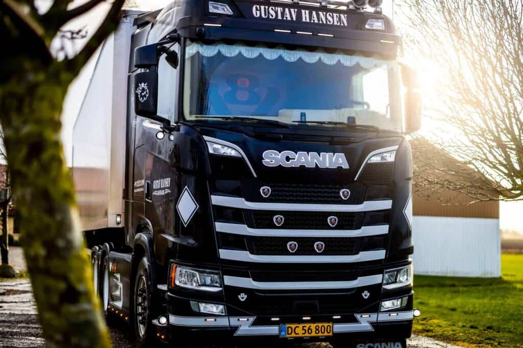 Fronten på en hvid Scania lastbil fra Gustav Hansen parkeret foran en gul bygning.