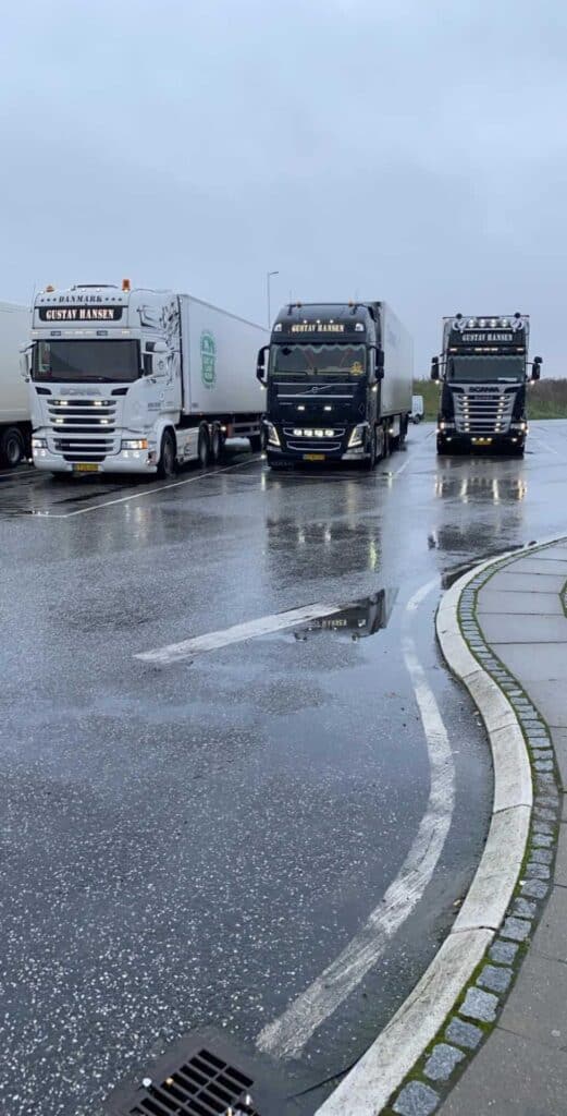 Tre store lastbiler fra vognmand Gustav Hansen holder på en våd parkeringsplads.