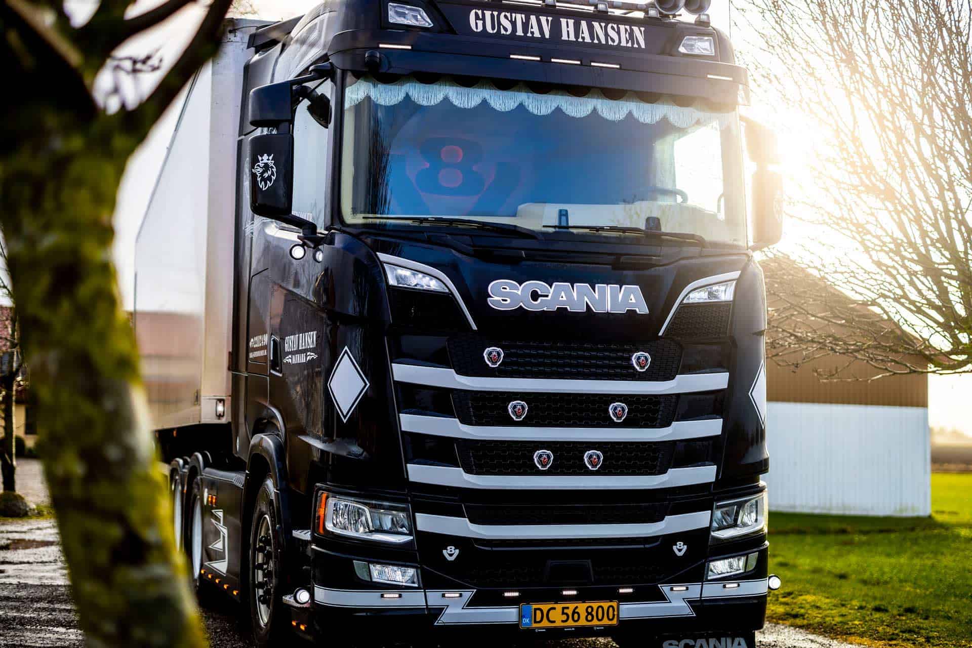 Fronten på en hvid Scania lastbil fra Gustav Hansen parkeret foran en gul bygning.