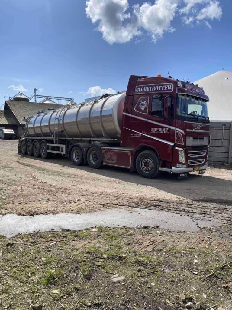 Mørkerød tankvogn fra Gustav Hansen transport parkeret foran en landbrugsbygning.