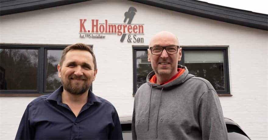 To glade repræsentanter for VVS-firmaet opstillet foran bygningens facadeskilt.