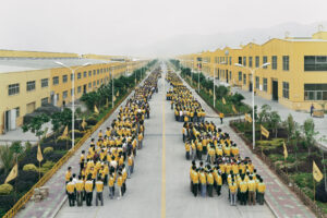Factory workers lined up in yellow uniforms in front of yellow buildings. Edward Burtynsky, China.