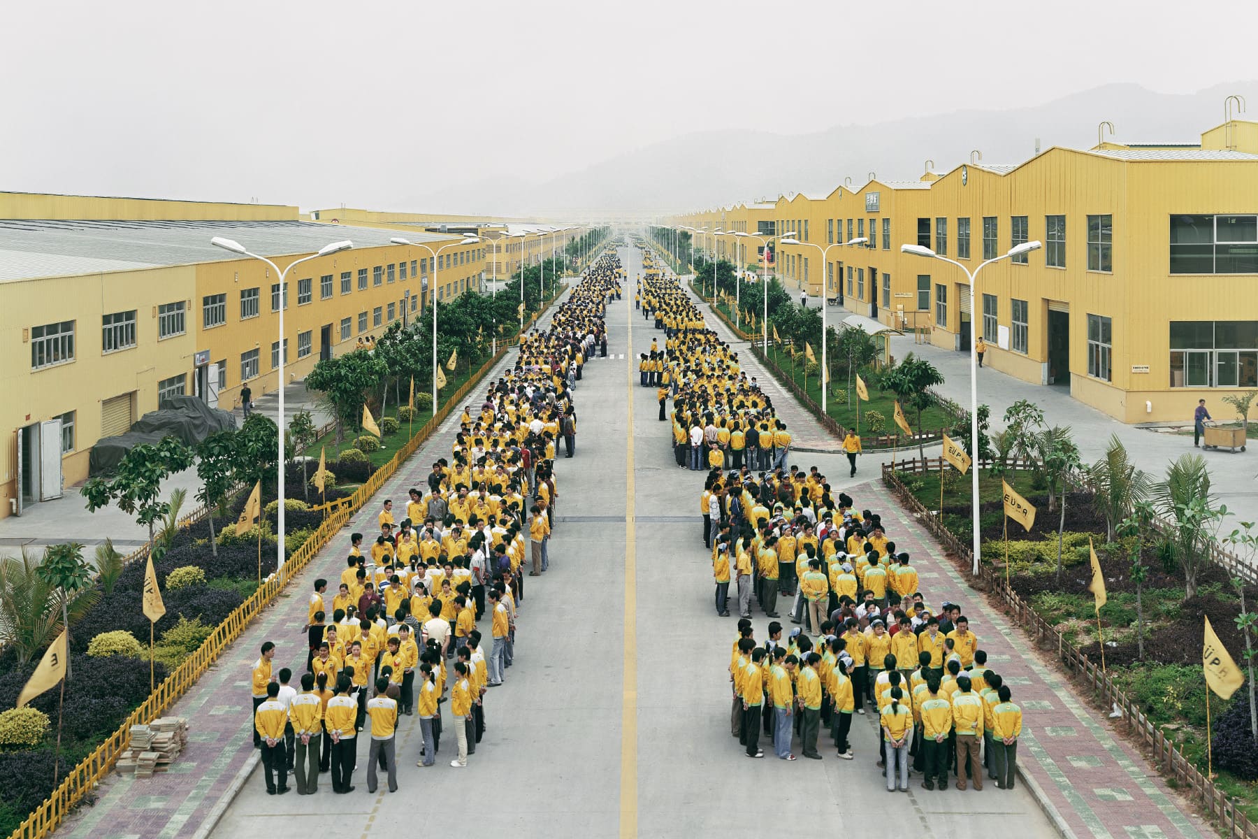 Factory workers lined up in yellow uniforms in front of yellow buildings. Edward Burtynsky, China.