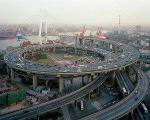 Wide city view featuring a bridge, industrial factories, and a spiraling freeway interchange. Edward Burtynsky, China.