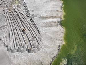 Overhead view of a lone machine piling gray material next to deep green water. Edward Burtynsky Anthropocene
