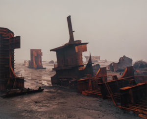 Rusting shipwreck pieces on muddy ground under overcast skies. Edward Burtynsky Shipbreaking