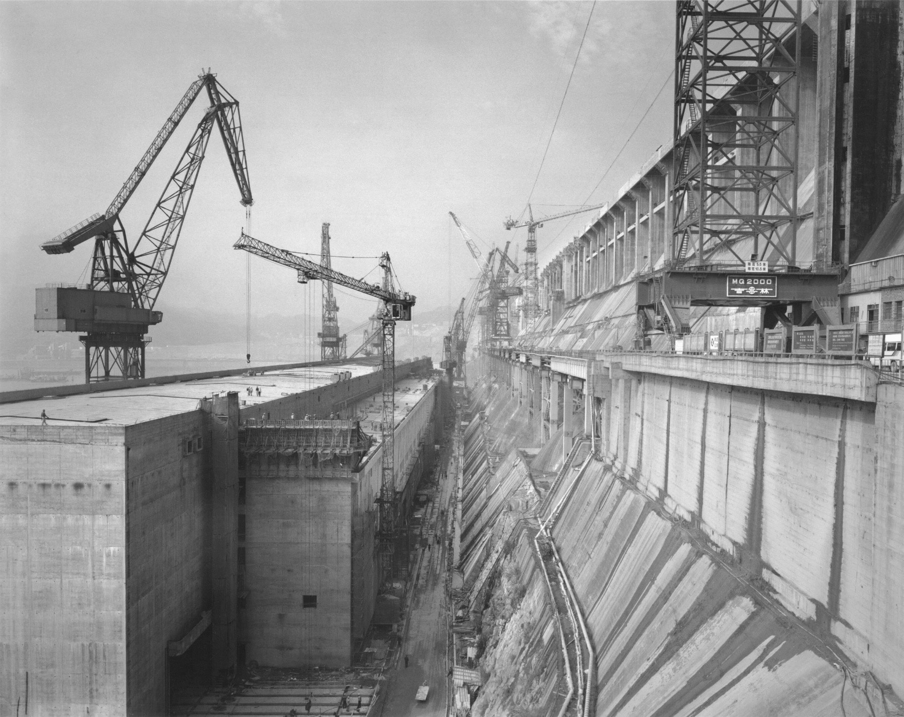 Black and white view of cranes and people alongside a dam. Edward Burtynsky The Source Before the Flood China