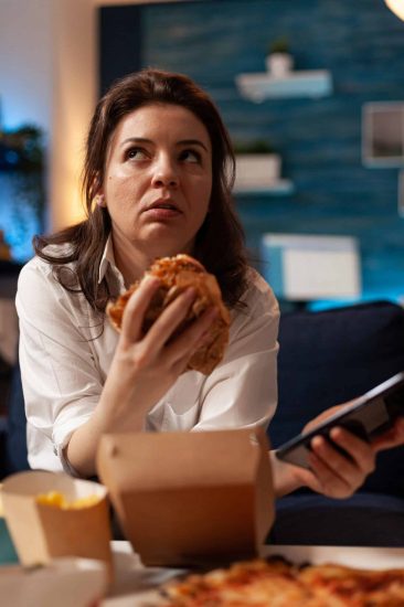 Caucasian female holding tastu burgery in hands browsing on social media using smartphone Caucasian female holding tastu burgery in hands browsing on social media using smartphone relaxing on couch during junck-food night. Woman enjoying takeaway food home delivery