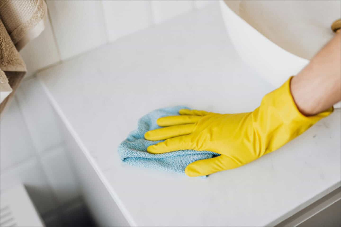 Close-up of a person wearing a yellow rubber glove wiping down a white marble bathroom countertop with a blue microfiber cloth.