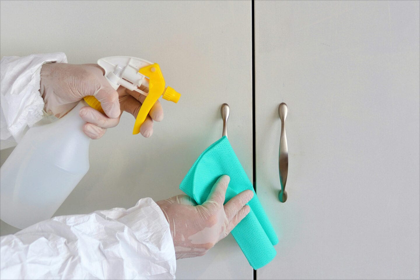 A person wearing a white protective suit and clear gloves holds a spray bottle and a green cloth to clean and sanitize the handles of a white cabinet.