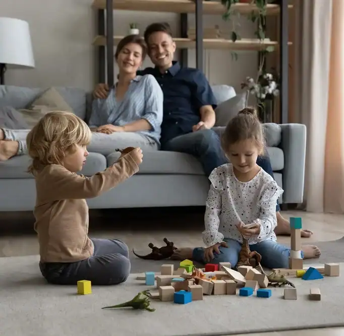 Two young children sit on a rug playing with wooden blocks and toy dinosaurs. A smiling adult couple sits on a couch in the background, watching the children. The room is cozy with soft lighting, a lamp, shelves, and houseplants.