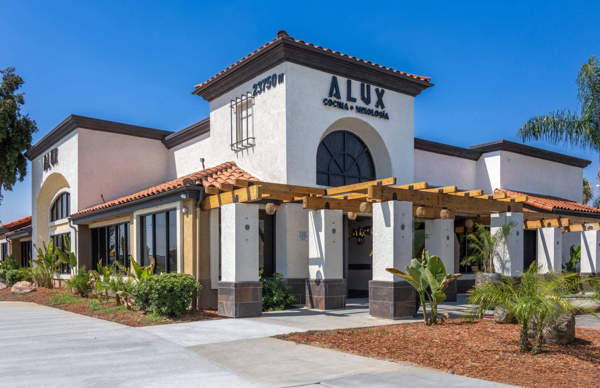 Alux Cocina y Vino restaurant in Moreno Valley, California, featuring a Mediterranean-style building with a terracotta roof, lush landscaping, and a welcoming entrance.