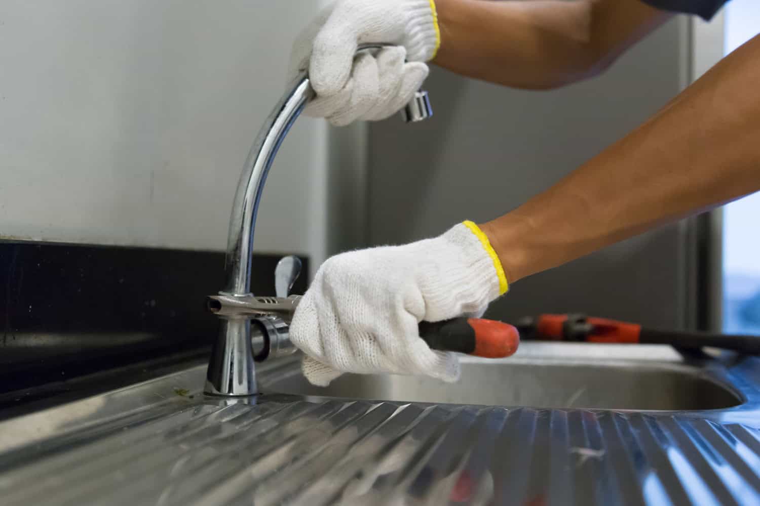 A man wearing utility gloves holds a wrench and works on a leaky kitchen sink faucet.