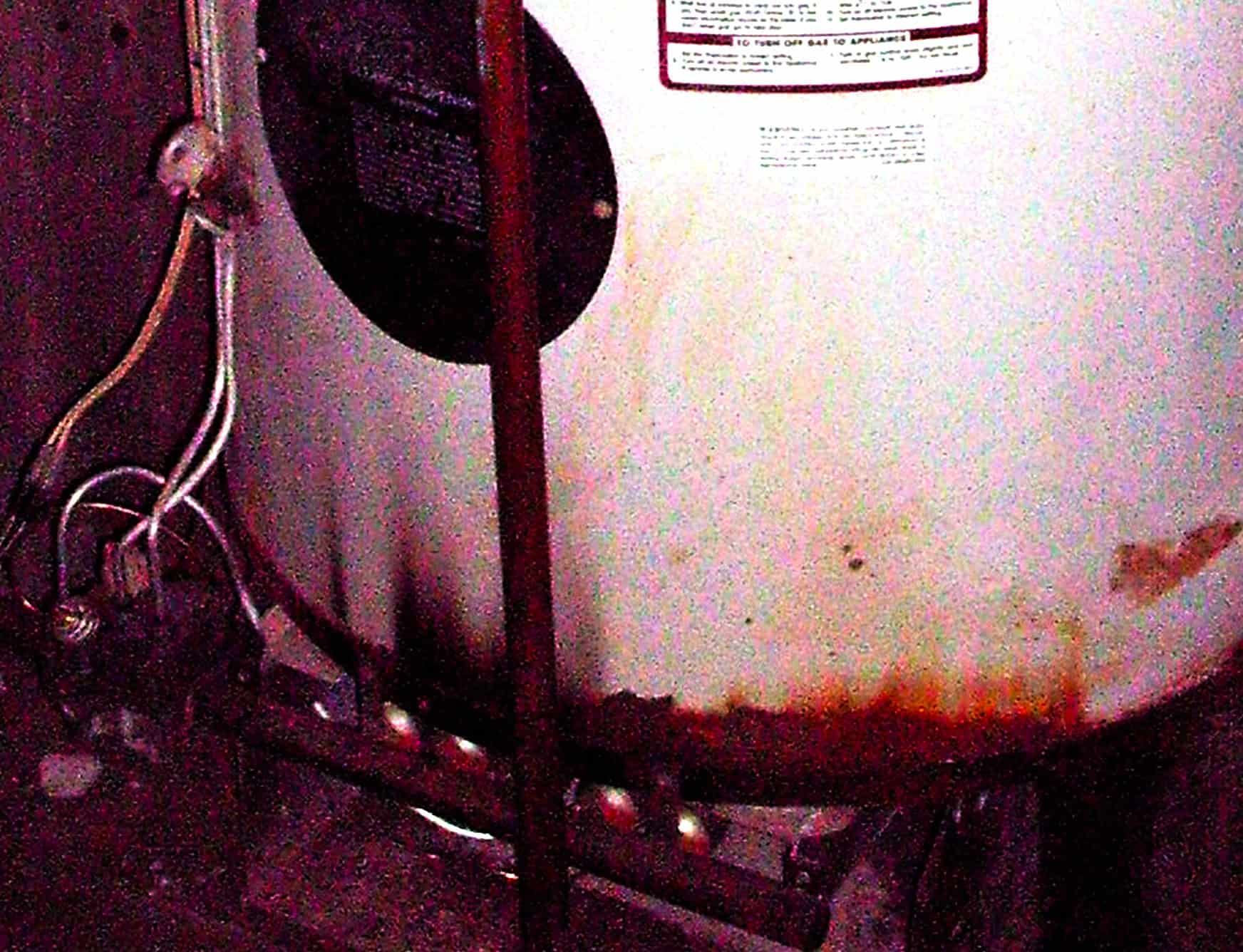 A slightly grainy image of the bottom of a white water heater, with chipped and rusted paint and flame burn marks along the bottom edge.
