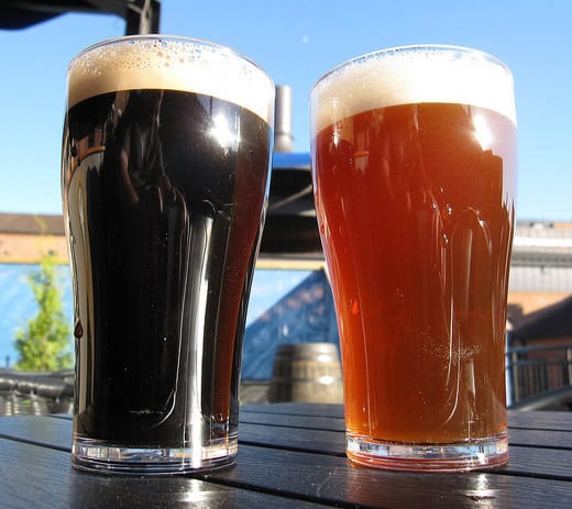 A pint of dark stout beer and a pint of light pale amber colored ale beer sit side by side on an outdoor picnic table.
