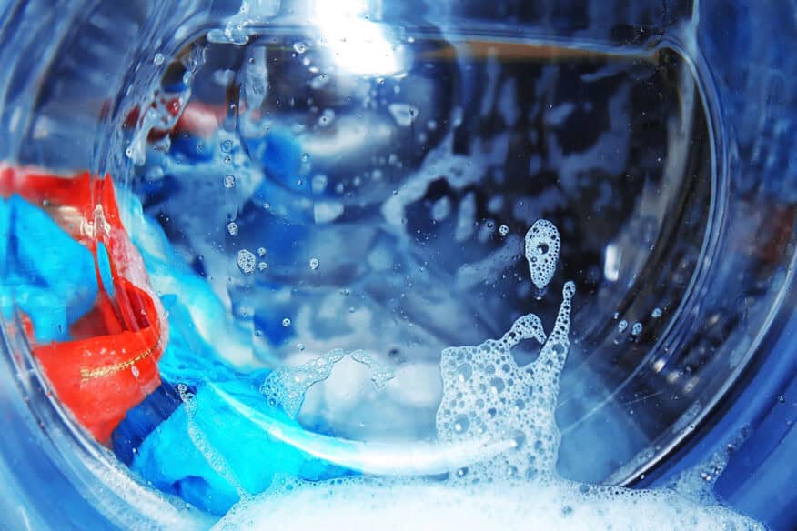A close-up of colorful clothes in a wet washing machine with soap bubbles dripping down the glass. Using less detergent is a benefit of a water softener.