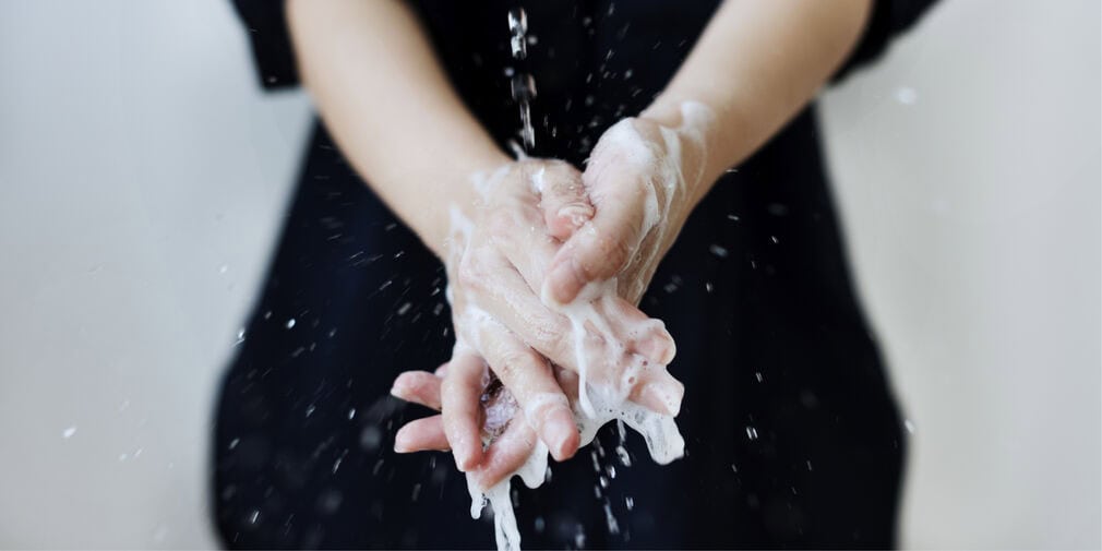 Soapy hands are in the foreground under a stream of water, while part of a woman in black is in the background. Restaurant hot water requirements need to be met to prevent scalding and burns. 