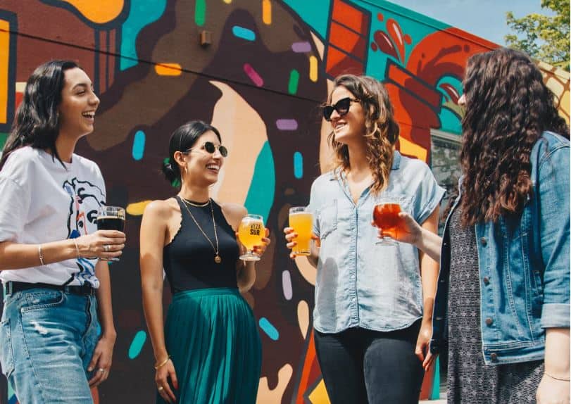 A group of four women stand together, holding pints of beer and talking in an outdoor beer garden. 