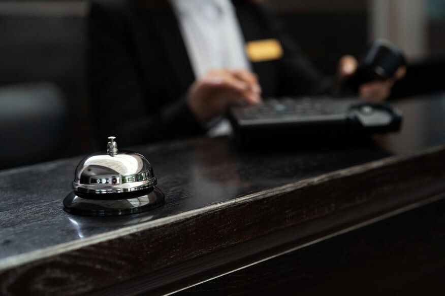 An old-fashioned bell on the front desk of a secure hotel, with a clerk in the background making a phone call.