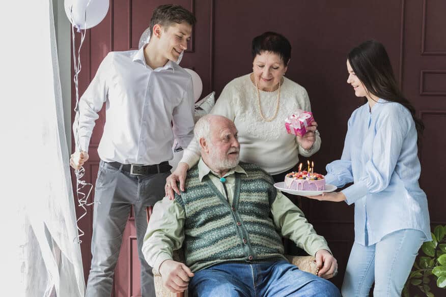 An elderly man sits in a chair in an assisted living facility while his family celebrates his birthday with balloons and cake.