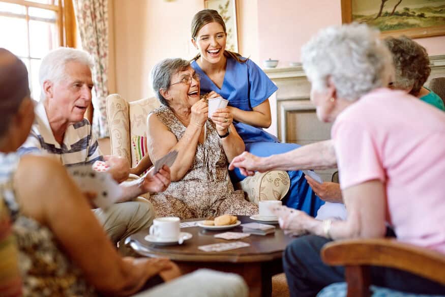 Elderly women and men play an exciting game of cards in an assisted living facility with a nurse smiling nearby.